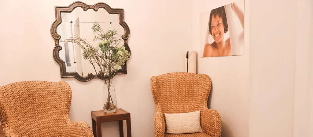 - Marion Salon Spa in Salt Point, NY Two wicker chairs with a pillow and a small wooden table with a vase of flowers sit beside a decorative mirror and a wall photo of a smiling woman, her look perfected by hand-tied hair extensions, in a softly lit room. - Marion Salon Spa in Salt Point, NY