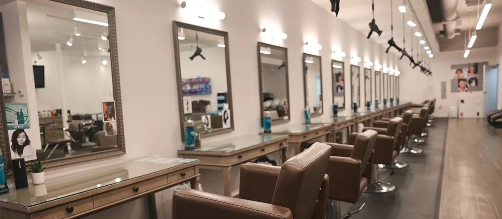 - Marion Salon Spa in Salt Point, NY A row of empty salon chairs and mirrors lines one wall of a modern, brightly lit hair salon with wooden floors and hairdryers hanging above, offering a welcoming space for clients seeking balayage hair color technique or luxurious hand-tied hair extensions. - Marion Salon Spa in Salt Point, NY