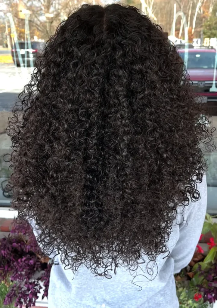 - Marion Salon Spa in Salt Point, NY Close-up of the back of a woman's full, voluminous dark curls photographed outdoors near flowers, highlighting the natural fullness enhanced by hand-tied hair extensions. - Marion Salon Spa in Salt Point, NY