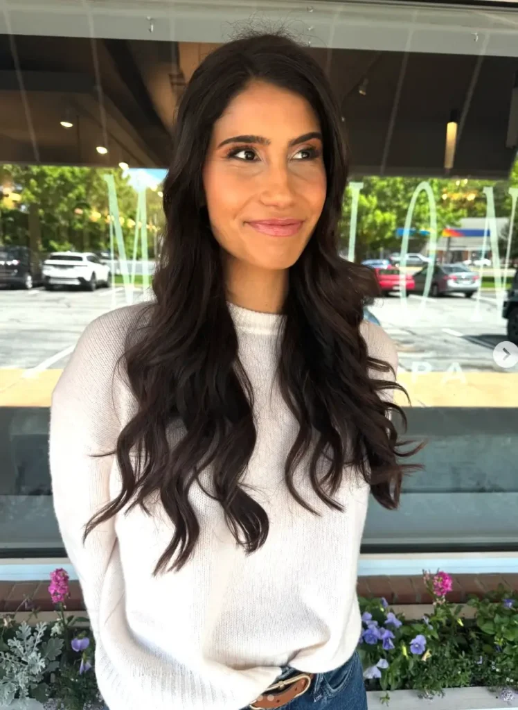 - Marion Salon Spa in Salt Point, NY A smiling woman with long, dark wavy hair and bold, defined lashes poses outdoors in a cream sweater near a flower bed, highlighting the fullness achieved with volume eyelash extensions. - Marion Salon Spa in Salt Point, NY