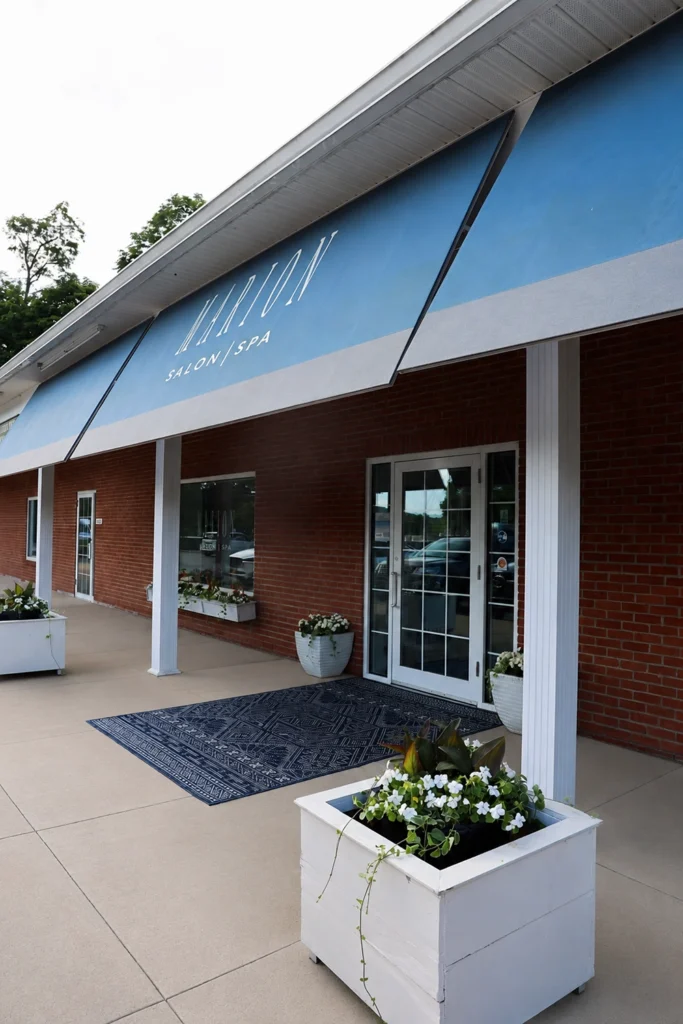 - Marion Salon Spa in Salt Point, NY Exterior of a salon/spa with blue awnings, glass entrance doors, white planters with flowers, and a black mat on a light-colored sidewalk. - Marion Salon Spa in Salt Point, NY